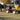 A woman and young girl sit on a bench seat of a red forecart, driving away from the camera. They’re pulling a large, yellow, cone-shaped seed spreader across a yard, while driving a team of two black Percheron draft horses.